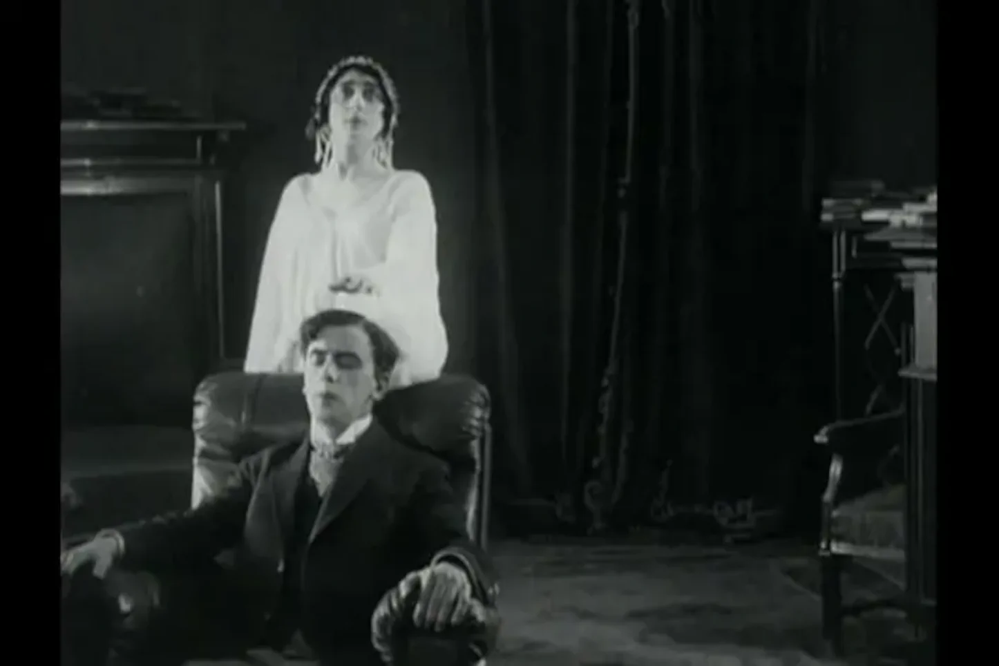 Still from Evgenii Bauer's 1915 silent film After Death, showing a man seated in an armchair while a ghostly female figure in white stands behind him in a darkened room
