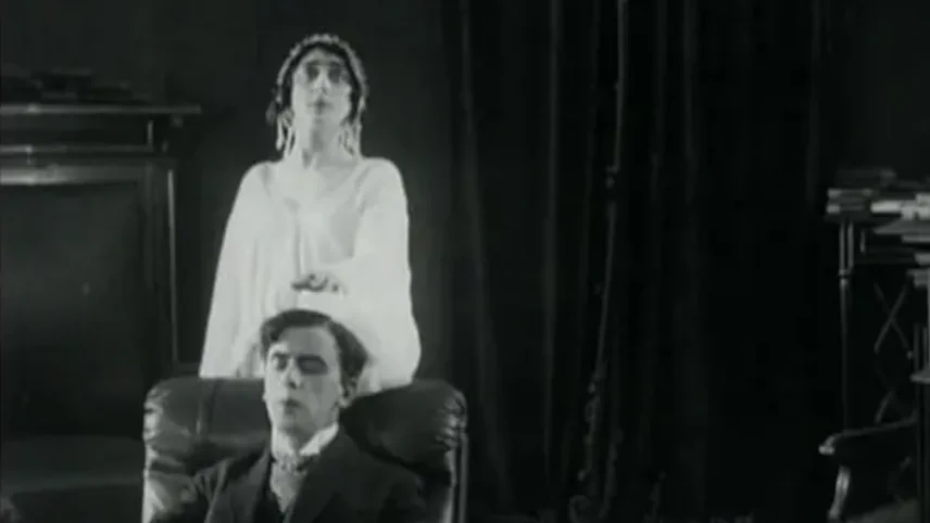 Still from a silent film: a man seated in an armchair while a ghostly female figure in white stands behind him in a darkened room