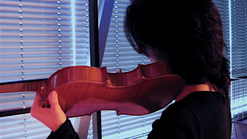 Monica Germino performing The Bravery of Women, facing away from the camera, holding a violin, silhouetted against vertical window blinds lit in blue and purple light