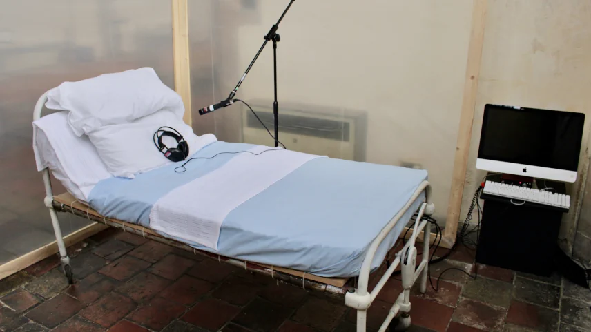 Installation view: a hospital bed with white and blue bedding, a microphone on a stand positioned above it, and a monitor on a bedside table.