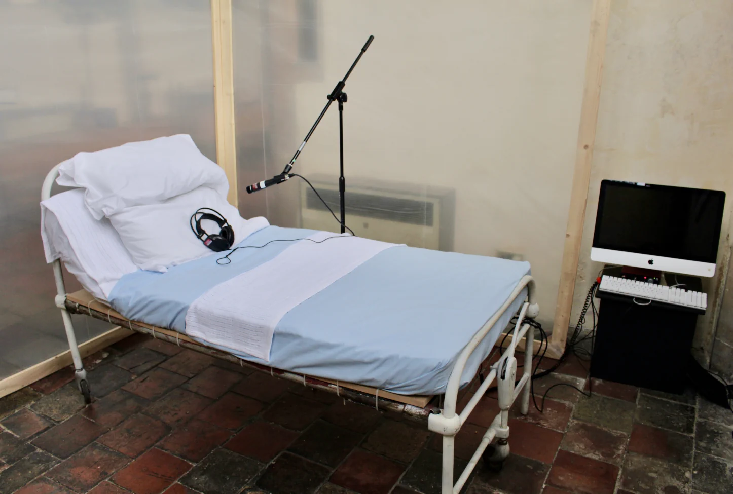 Installation view: a hospital bed with white and blue bedding, a microphone on a stand positioned above it, and a monitor on a bedside table.