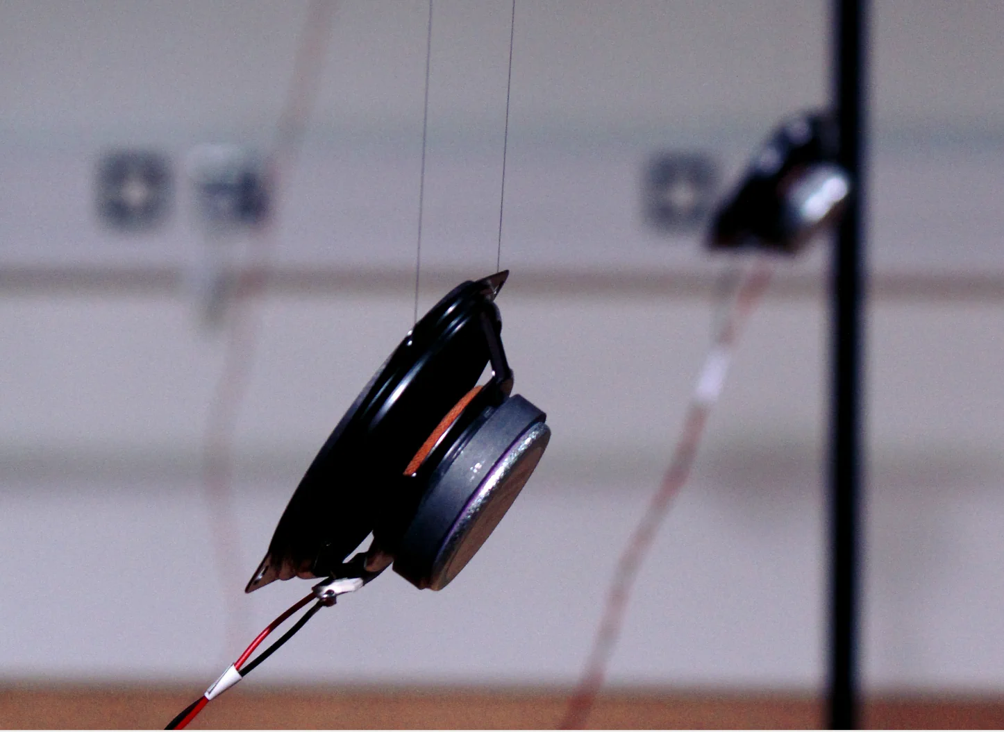 Close-up of a small loudspeaker suspended by wires with red and black connecting cables, against a blurred background