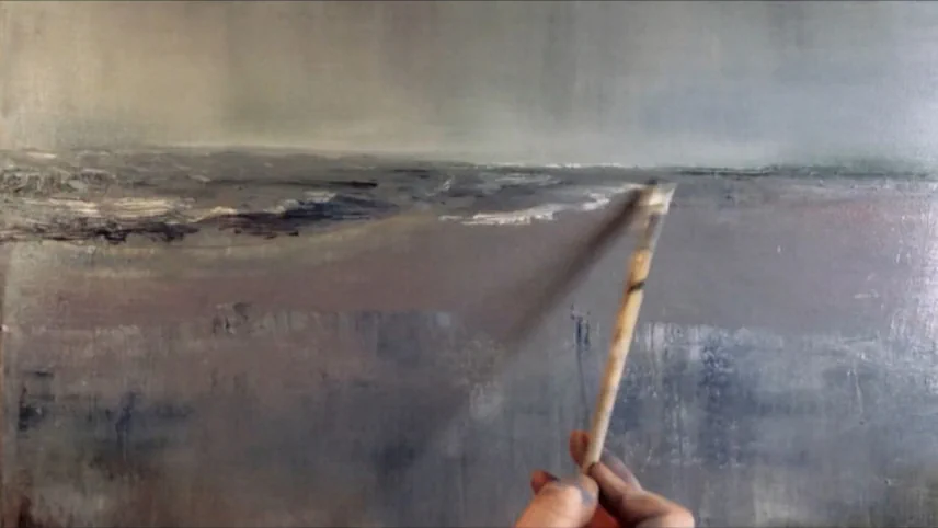 A hand holding a paintbrush against a canvas showing a muted seascape with a hazy horizon, the painting in progress