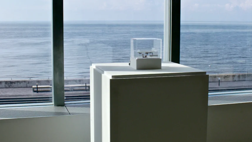Installation view: a small object in a transparent display case on a white plinth, positioned in front of large windows overlooking the sea
