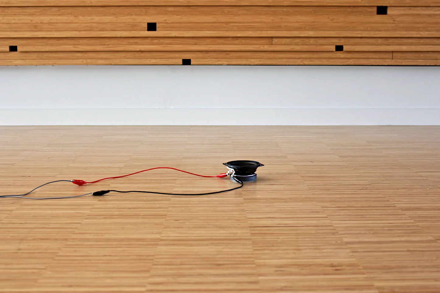 Installation view: a small loudspeaker with red and black wires lying on a light wood table in front of a slatted wood panel with small black square markers