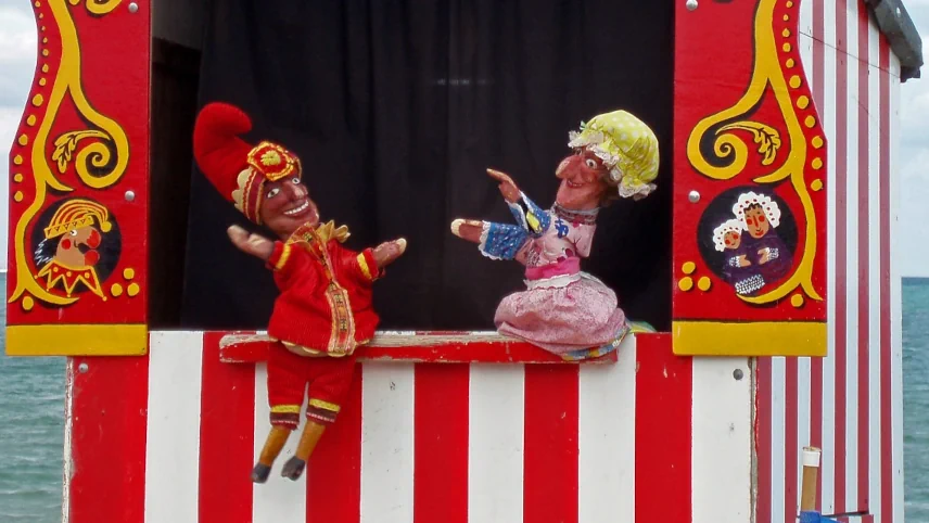 A traditional Punch and Judy puppet show booth with red and white striped front, showing Mr Punch in red costume and a female puppet facing each other on the ledge, with a painted decorative arch above
